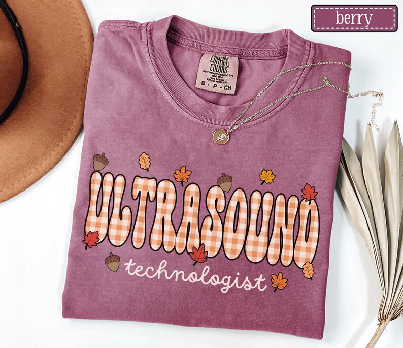 Puede incluir: Camiseta color bayas con la frase "ULTRASOUND technologist" en un estilo retro con un estampado de cuadros. El dise&ntilde;o incluye hojas de oto&ntilde;o y bellotas. La camiseta tiene cuello redondo y mangas cortas. Un collar dorado est&aacute; sobre la camiseta.