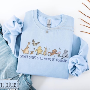 May include: Pale blue sweatshirt with a cartoon character design and the words "SMALL STEPS STILL MOVE US FORWARD." The sweatshirt is shown with blue jeans. A silver necklace is draped on the sweatshirt.