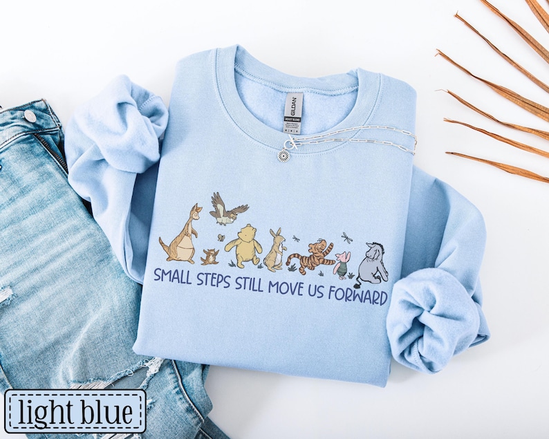 May include: Pale blue sweatshirt with a cartoon character design and the words "SMALL STEPS STILL MOVE US FORWARD." The sweatshirt is shown with blue jeans. A silver necklace is draped on the sweatshirt.