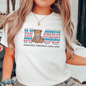 Puede incluir: Camiseta blanca con un gr&aacute;fico a rayas rosa, azul y blanco que dice "NICU" con un oso de peluche marr&oacute;n en el medio. El texto debajo del gr&aacute;fico dice "unidad de cuidados intensivos neonatales".
