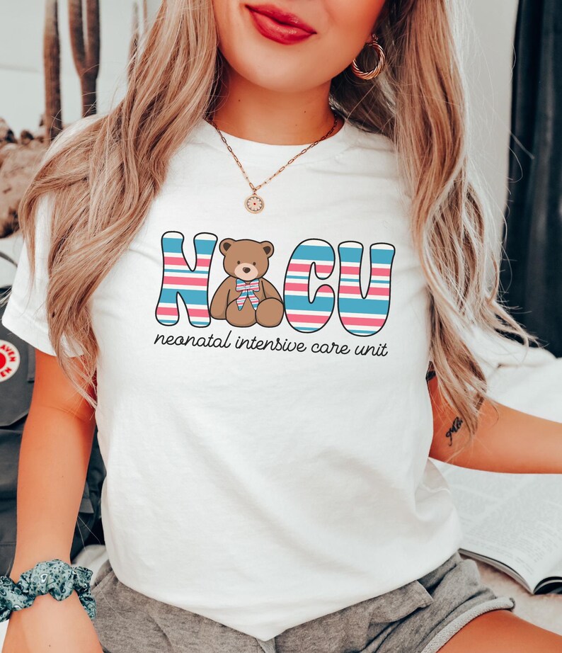 Puede incluir: Camiseta blanca con un gr&aacute;fico a rayas rosa, azul y blanco que dice "NICU" con un oso de peluche marr&oacute;n en el medio. El texto debajo del gr&aacute;fico dice "unidad de cuidados intensivos neonatales".