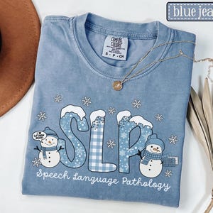 May include: Blue jean Comfort Colors t-shirt with a winter-themed design. The shirt features the letters "SLP" in blue and white, with snow-capped letters and snowflakes. The words "Speech Language Pathology" are printed below.