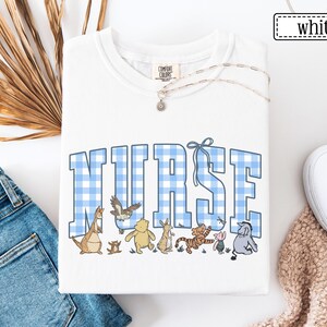 K&ouml;nnte beinhalten: Wei&szlig;es Sweatshirt mit Rundhalsausschnitt und dem Wort &bdquo;NURSE&ldquo; in blau-wei&szlig; karierten Buchstaben. Das Sweatshirt zeigt Zeichentrickfiguren unter dem Wort &bdquo;NURSE&ldquo;. Eine silberne Halskette mit einem kleinen Anh&auml;nger ist sichtbar. Das Wort &bdquo;wei&szlig;&ldquo; befindet sich in einem wei&szlig;en Rechteck.