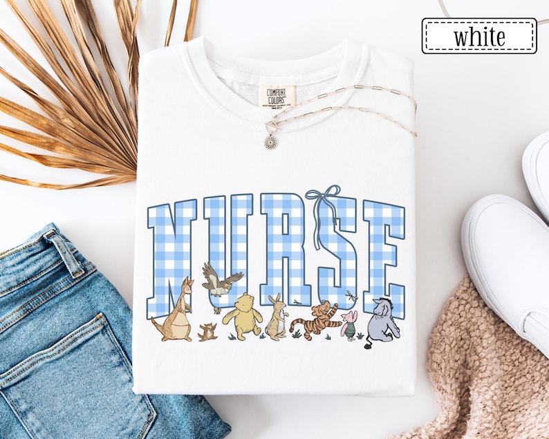 K&ouml;nnte beinhalten: Wei&szlig;es Sweatshirt mit Rundhalsausschnitt und dem Wort &bdquo;NURSE&ldquo; in blau-wei&szlig; karierten Buchstaben. Das Sweatshirt zeigt Zeichentrickfiguren unter dem Wort &bdquo;NURSE&ldquo;. Eine silberne Halskette mit einem kleinen Anh&auml;nger ist sichtbar. Das Wort &bdquo;wei&szlig;&ldquo; befindet sich in einem wei&szlig;en Rechteck.