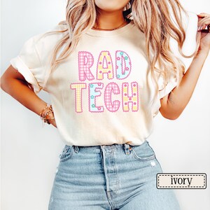 May include: Ivory-colored t-shirt with the text "RAD TECH" in a colorful, patterned design. The letters are outlined in a contrasting color and filled with gingham and star patterns. The shirt is a casual style.