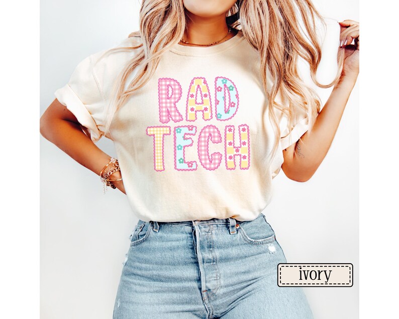May include: Ivory-colored t-shirt with the text "RAD TECH" in a colorful, patterned design. The letters are outlined in a contrasting color and filled with gingham and star patterns. The shirt is a casual style.