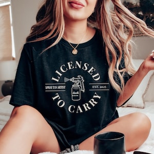 May include: Black t-shirt with a white circle graphic that says "Licensed to Carry" with a spray tan gun and bottle of "dark" tan. The text also says "Spray Tan Artist" and "Est. 2025".