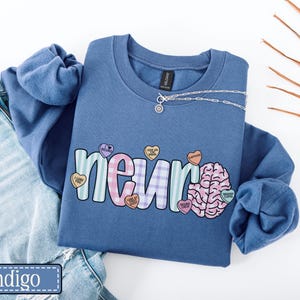 May include: Indigo blue sweatshirt with the word "neuro" in a colorful, playful font, with a brain graphic and heart-shaped candies. A silver necklace is draped across the top. The sweatshirt is paired with blue jeans.