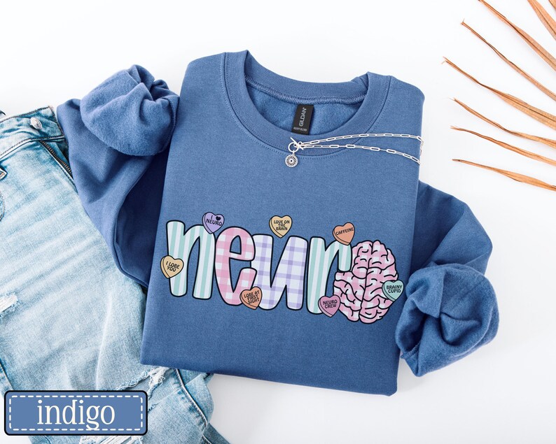 May include: Indigo blue sweatshirt with the word "neuro" in a colorful, playful font, with a brain graphic and heart-shaped candies. A silver necklace is draped across the top. The sweatshirt is paired with blue jeans.