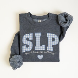 May include: A dark gray sweatshirt with a blue and white checkered "SLP" graphic and the text "speech language pathology" below. A small white heart is centered below the text.