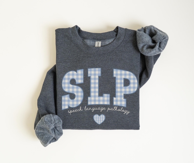 May include: A dark gray sweatshirt with a blue and white checkered "SLP" graphic and the text "speech language pathology" below. A small white heart is centered below the text.