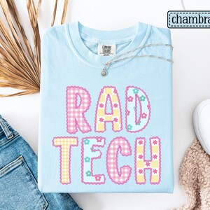 May include: Light blue t-shirt with the words "RAD TECH" in a colorful, gingham-style design. The letters are outlined with a floral pattern. A silver necklace and white shoes are also visible.