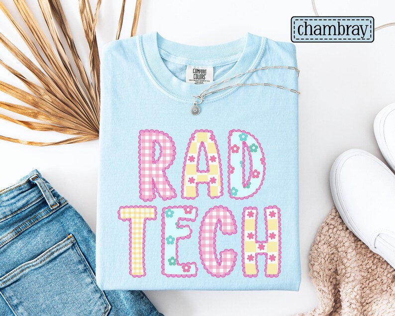 May include: Light blue t-shirt with the words "RAD TECH" in a colorful, gingham-style design. The letters are outlined with a floral pattern. A silver necklace and white shoes are also visible.