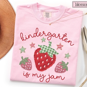 May include: A light pink t-shirt featuring a central strawberry applique with a green gingham leaf, flanked by two smaller gingham strawberries. The text reads kindergarten is my jam in red script, accented by small red and green gingham stars.
