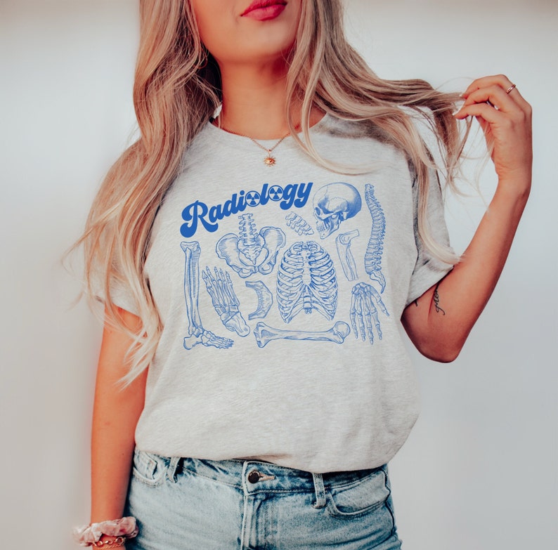 May include: A light gray t-shirt with the word "Radiology" in blue script, accompanied by blue illustrations of human skeletal anatomy. The design includes a skull, spine, rib cage, and various bones.