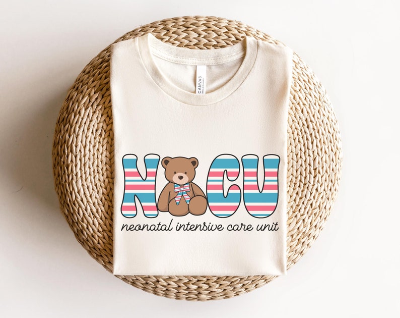 Puede incluir: Una camiseta de color crema con un gr&aacute;fico de un osito de peluche en el centro de las letras "NICU" en rayas azules, rosas y blancas. El texto "unidad de cuidados intensivos neonatales" est&aacute; debajo del gr&aacute;fico.