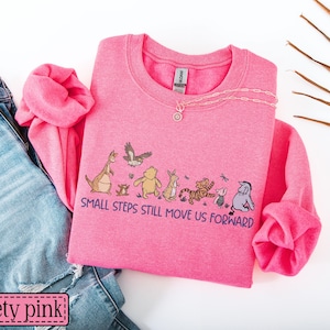 May include: A safety pink sweatshirt featuring the text "SMALL STEPS STILL MOVE US FORWARD" and a cartoon character graphic. The sweatshirt is styled with blue jeans. A gold necklace is placed on the sweatshirt.