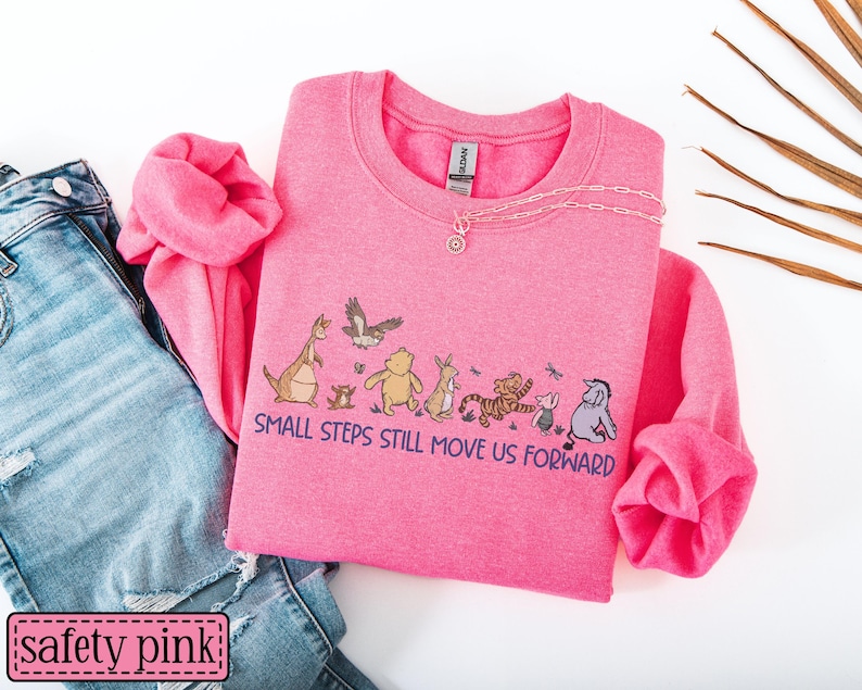 May include: A safety pink sweatshirt featuring the text "SMALL STEPS STILL MOVE US FORWARD" and a cartoon character graphic. The sweatshirt is styled with blue jeans. A gold necklace is placed on the sweatshirt.