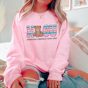 Puede incluir: Sudadera rosa con un gr&aacute;fico de un osito de peluche y el texto "NICU unidad de cuidados intensivos neonatales" en rayas azules, rojas y blancas.