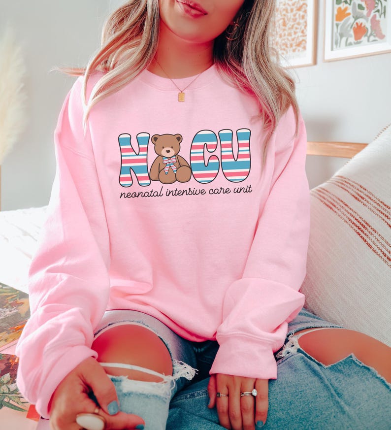 Puede incluir: Sudadera rosa con un gr&aacute;fico de un osito de peluche y el texto "NICU unidad de cuidados intensivos neonatales" en rayas azules, rojas y blancas.
