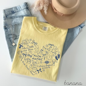 May include: A yellow t-shirt with a heart-shaped design in blue, featuring the words "NICU nurse" and other related phrases and illustrations. The shirt is paired with light blue jeans, a tan hat, and a necklace.