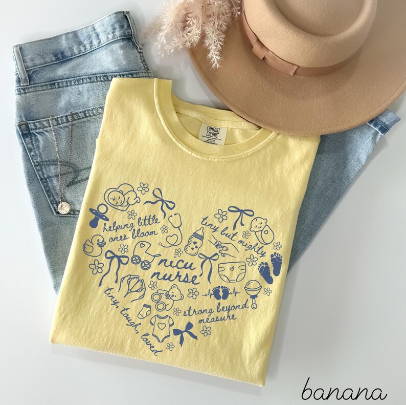 May include: A yellow t-shirt with a heart-shaped design in blue, featuring the words "NICU nurse" and other related phrases and illustrations. The shirt is paired with light blue jeans, a tan hat, and a necklace.