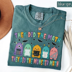 May include: A blue-green t-shirt featuring a graphic of five colorful monsters holding math tools. The text reads "THEY DID THE MATH" and "THEY DID THE MONSTER MATH" in a bubbly, multi-colored font. Small black bats and stars are scattered around the monsters.