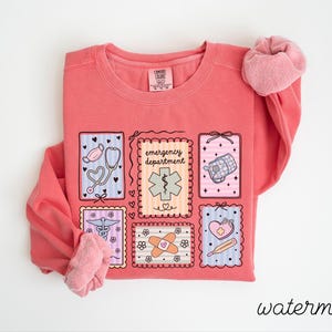 Pode incluir: Uma camisola coral com gola redonda e mangas compridas. A frente apresenta um design de seis ilustra&ccedil;&otilde;es emolduradas de s&iacute;mbolos m&eacute;dicos, incluindo um estetosc&oacute;pio, uma mala m&eacute;dica e um penso. A palavra "emergency department" tamb&eacute;m &eacute; exibida.