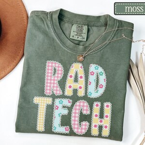 May include: A moss green t-shirt with the words "RAD TECH" in a colorful, patterned design. The letters are decorated with pink gingham, yellow gingham, and blue floral patterns. A gold necklace rests on the shirt.
