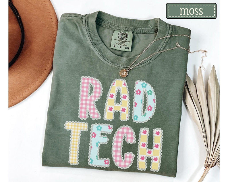 May include: A moss green t-shirt with the words "RAD TECH" in a colorful, patterned design. The letters are decorated with pink gingham, yellow gingham, and blue floral patterns. A gold necklace rests on the shirt.