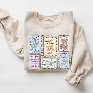May include: A beige sweatshirt with a colorful graphic design featuring medical-themed illustrations. The design includes a stethoscope, a teddy bear, a bandage, and the words "pediatric nurse" and "it's a good day to care for tiny humans."
