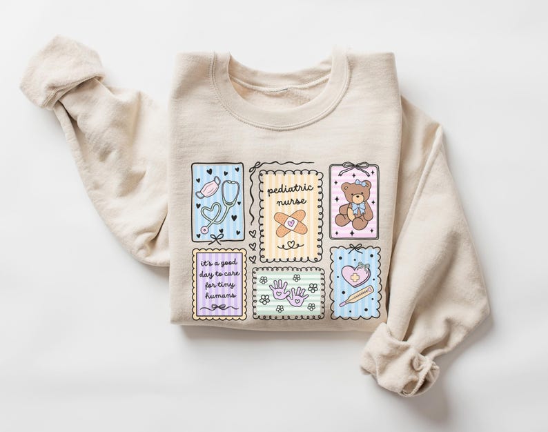 May include: A beige sweatshirt with a colorful graphic design featuring medical-themed illustrations. The design includes a stethoscope, a teddy bear, a bandage, and the words "pediatric nurse" and "it's a good day to care for tiny humans."
