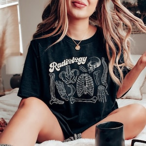 May include: Black t-shirt with the word "Radiology" in a white, retro font. The shirt features a detailed white illustration of human skeletal anatomy, including bones and the spine. The shirt is a relaxed fit.