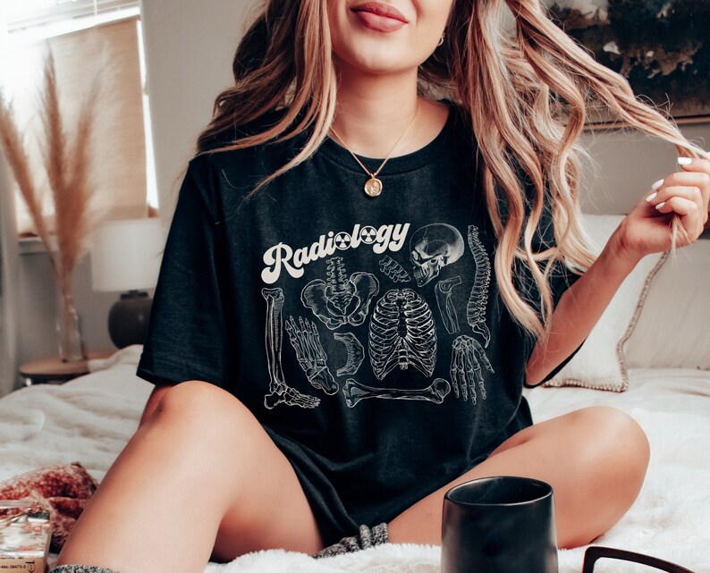 May include: Black t-shirt with the word "Radiology" in a white, retro font. The shirt features a detailed white illustration of human skeletal anatomy, including bones and the spine. The shirt is a relaxed fit.