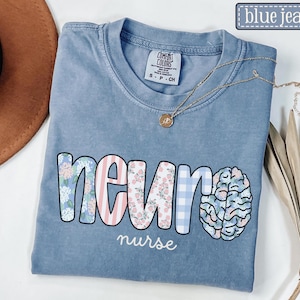 May include: Blue jean colored t-shirt with the word "NEURON" in a floral and striped design, and a brain graphic. The word "nurse" is written below. A gold necklace with a pendant is draped across the shirt.