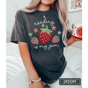 May include: A charcoal grey short-sleeve t-shirt featuring a graphic of three strawberries with green and red gingham patterns. The text "reading is my jam" is printed in white script above and below the strawberries, accented by small stars.