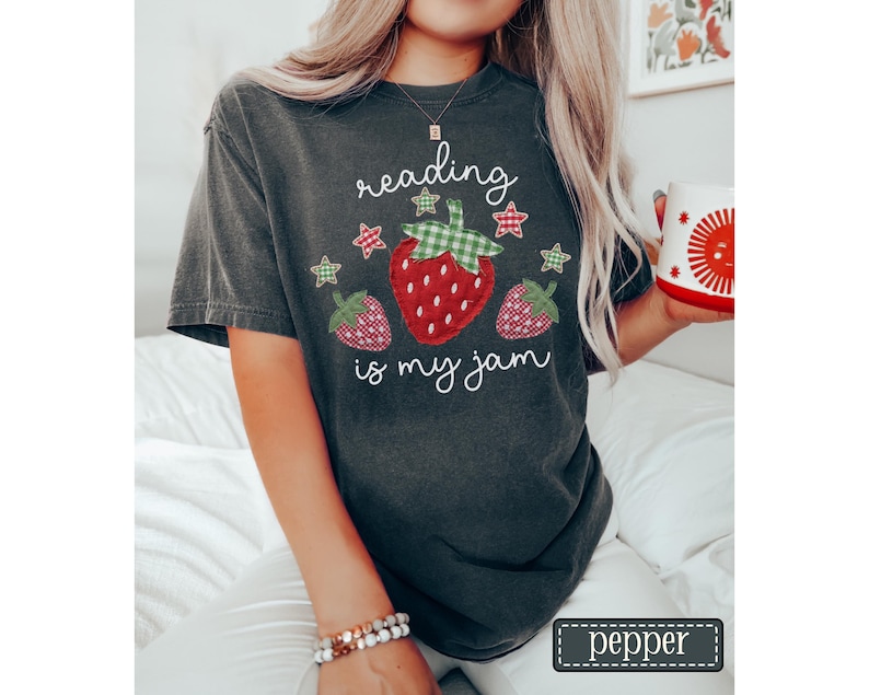 May include: A charcoal grey short-sleeve t-shirt featuring a graphic of three strawberries with green and red gingham patterns. The text "reading is my jam" is printed in white script above and below the strawberries, accented by small stars.
