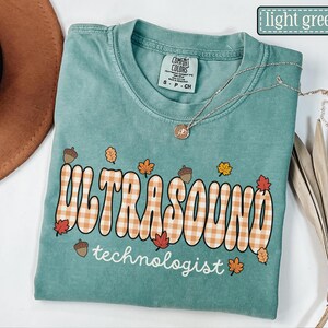 Puede incluir: Camiseta verde claro con la frase "ULTRASOUND technologist" en una fuente retro con un patr&oacute;n de cuadros, decorada con hojas de oto&ntilde;o y bellotas. Un collar plateado con un peque&ntilde;o colgante est&aacute; sobre la camiseta.