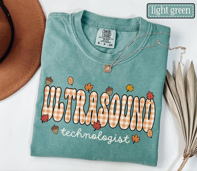 Puede incluir: Camiseta verde claro con la frase "ULTRASOUND technologist" en una fuente retro con un patr&oacute;n de cuadros, decorada con hojas de oto&ntilde;o y bellotas. Un collar plateado con un peque&ntilde;o colgante est&aacute; sobre la camiseta.