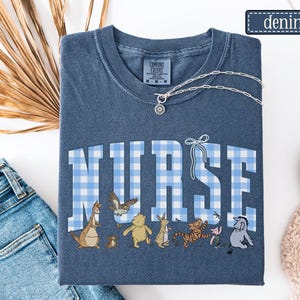 K&ouml;nnte beinhalten: Denimblaues T-Shirt mit dem Wort "NURSE" in gro&szlig;en, hellblau-wei&szlig; karierten Buchstaben. Unter dem Wort befinden sich Zeichentrickfiguren. Eine silberne Halskette mit einem kreisf&ouml;rmigen Anh&auml;nger liegt auf dem Shirt. Das Wort "Denim" steht in einem kleinen Rechteck.