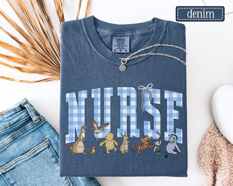 K&ouml;nnte beinhalten: Denimblaues T-Shirt mit dem Wort "NURSE" in gro&szlig;en, hellblau-wei&szlig; karierten Buchstaben. Unter dem Wort befinden sich Zeichentrickfiguren. Eine silberne Halskette mit einem kreisf&ouml;rmigen Anh&auml;nger liegt auf dem Shirt. Das Wort "Denim" steht in einem kleinen Rechteck.