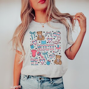 May include: A gray t-shirt with a colorful design featuring the word "NICU" surrounded by baby-themed icons like teddy bears, hearts, pacifiers, and footprints.