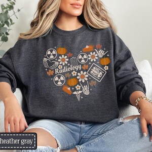 May include: A dark heather gray crewneck sweatshirt featuring a heart-shaped graphic composed of radiology symbols, pumpkins, autumn leaves, flowers, a skeleton hand, and a medical caduceus. The word Radiology is printed in the center of the heart.