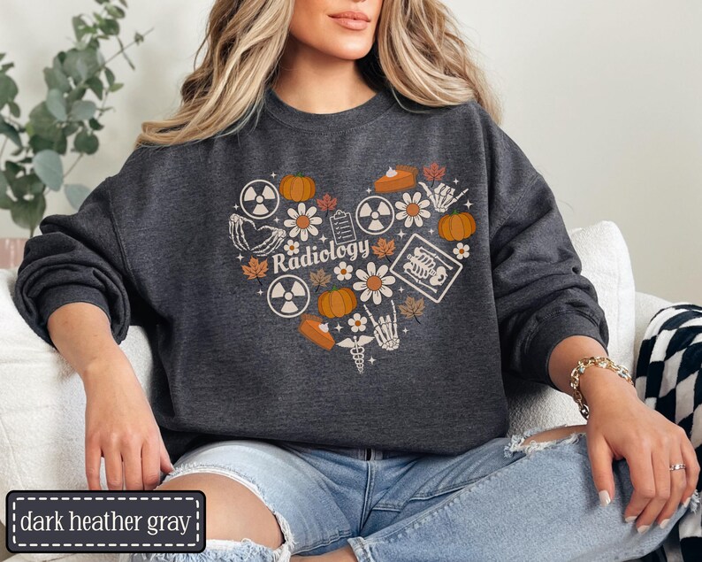 May include: A dark heather gray crewneck sweatshirt featuring a heart-shaped graphic composed of radiology symbols, pumpkins, autumn leaves, flowers, a skeleton hand, and a medical caduceus. The word Radiology is printed in the center of the heart.