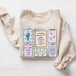 May include: A beige sweatshirt featuring several colorful rectangular designs with medical-themed illustrations. The designs include a skeleton hand, a radiology symbol, and the text "It's a good day to xray."
