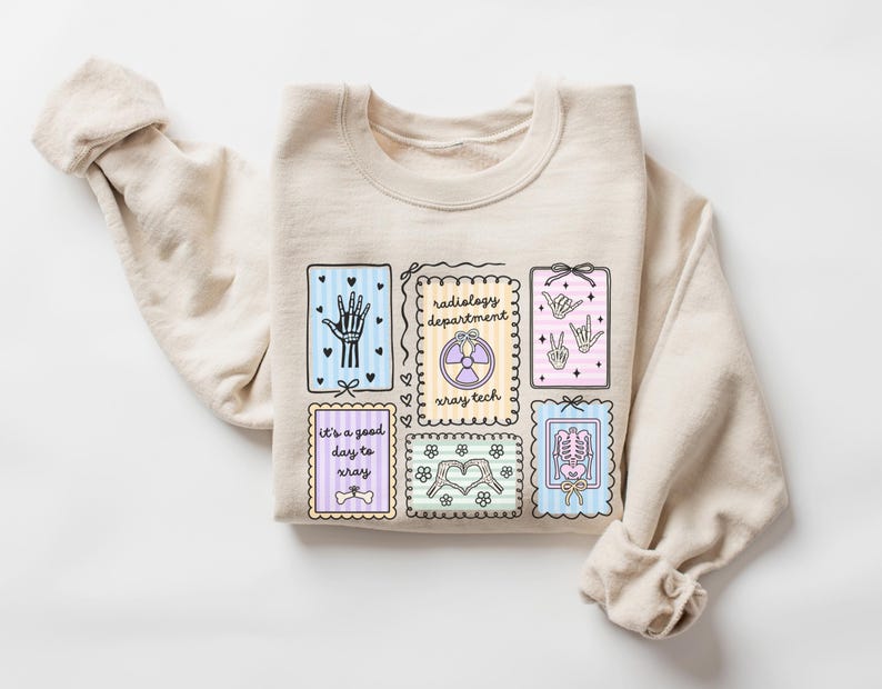 May include: A beige sweatshirt featuring several colorful rectangular designs with medical-themed illustrations. The designs include a skeleton hand, a radiology symbol, and the text "It's a good day to xray."