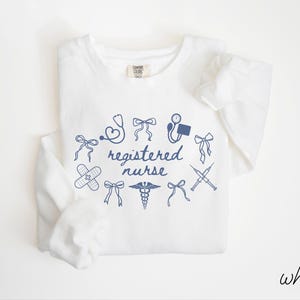 May include: White long-sleeve sweatshirt with a navy blue graphic design. The design includes medical symbols such as a stethoscope, band-aid, and caduceus, along with the words "registered nurse". The word "white" is written in the bottom right corner.