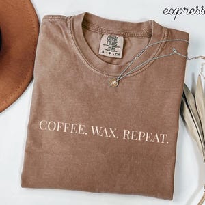 May include: A brown t-shirt with the text "COFFEE. WAX. REPEAT." in white. A silver necklace with a pendant is draped across the shirt. A brown hat and dried plant are in the background.