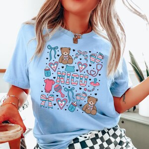 May include: Light blue t-shirt with a graphic design of baby-related icons and the word "NICU" in pink, blue, and white stripes. The icons include teddy bears, hearts, bows, pacifiers, baby bottles, and footprints.