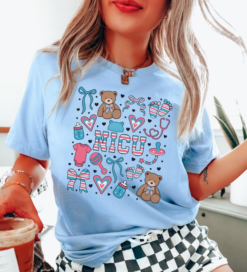 May include: Light blue t-shirt with a graphic design of baby-related icons and the word "NICU" in pink, blue, and white stripes. The icons include teddy bears, hearts, bows, pacifiers, baby bottles, and footprints.